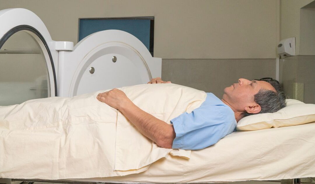 A man lies on a medical examination table, covered with a sheet, inside a circular scanning machine in a clinical setting.