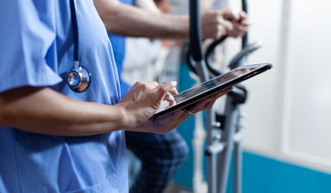 A healthcare professional in scrubs uses a tablet, standing beside gym equipment.