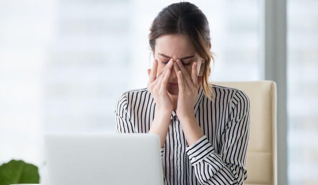 A woman in a striped shirt sits at a desk, rubbing her eyes in front of a laptop.